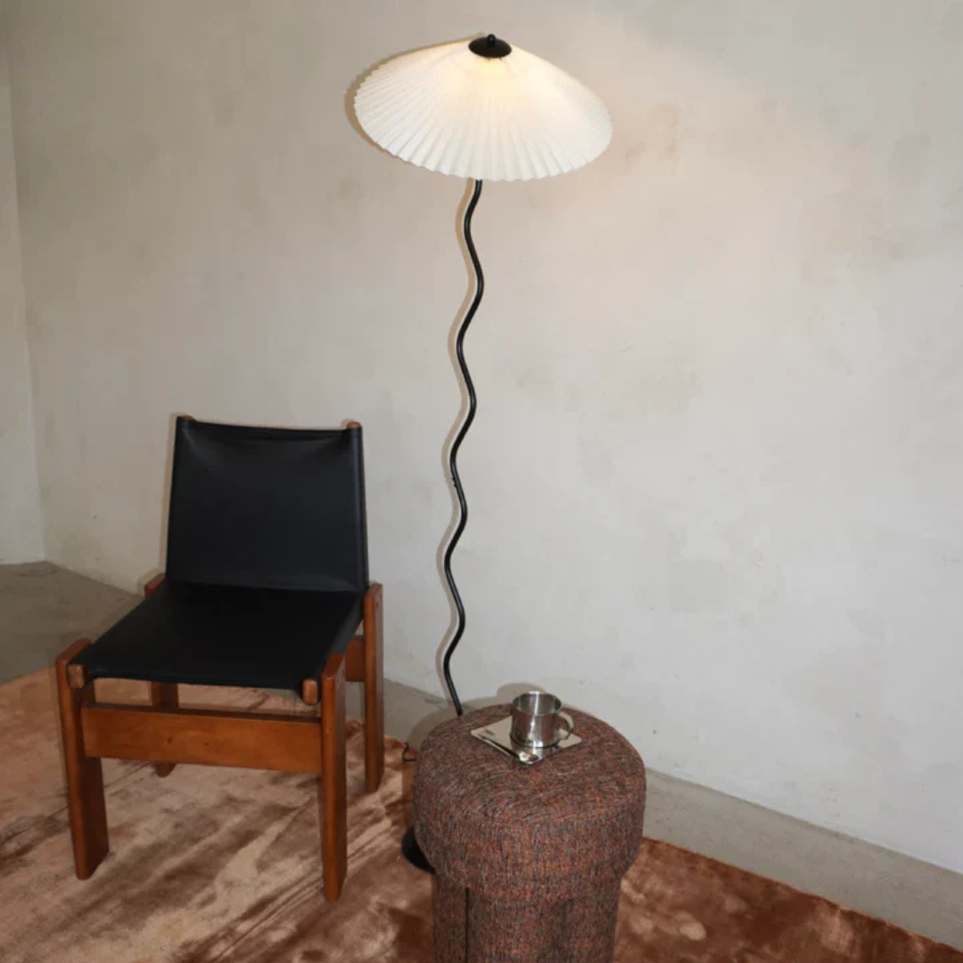 A modern lounge setup featuring the Seashell floor lamp with a wavy stand and pleated shade, paired with a wooden-framed leather chair, a textured ottoman serving as a side table, and a warm-toned rug against a neutral wall.