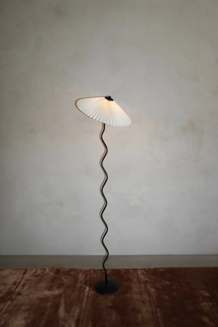 The Seashell floor lamp featuring a pleated shade and sculptural wavy stand, standing elegantly on a soft brown rug against a minimalist neutral wall, perfect for modern interiors.