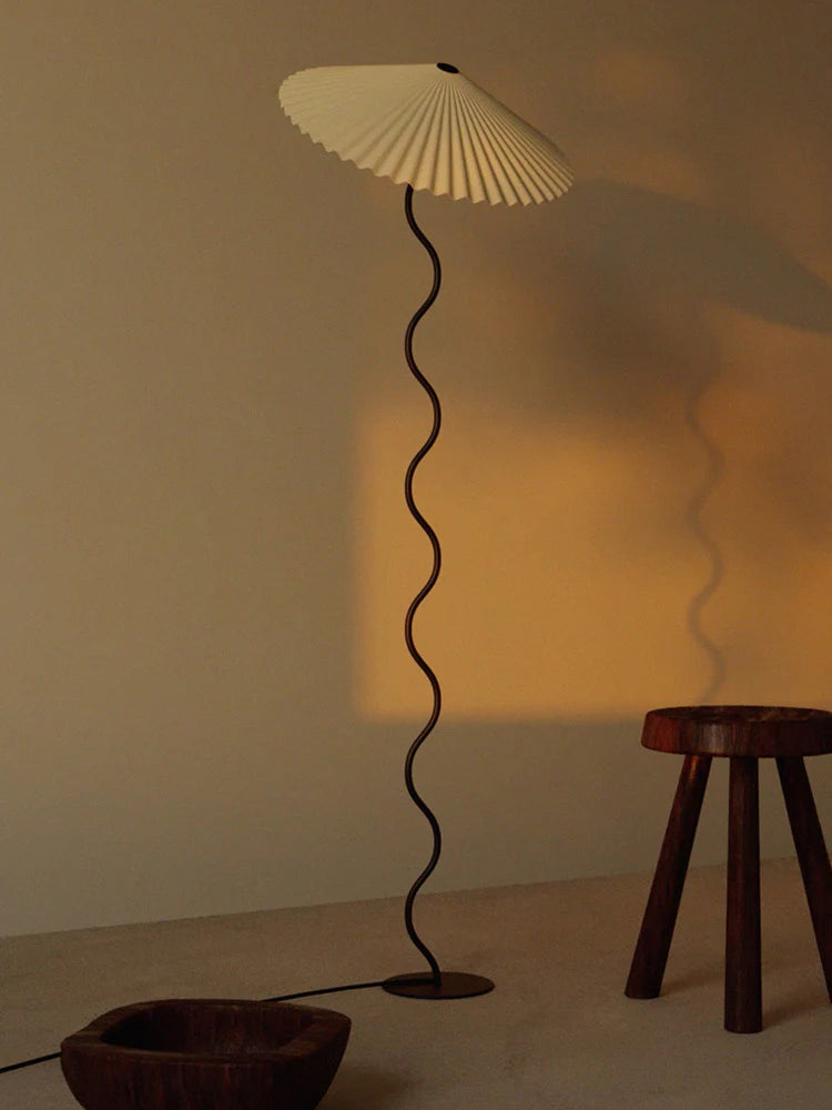 The Seashell floor lamp with a wavy stand and pleated shade, casting a warm glow against a neutral wall during sunset, paired with rustic wooden stools and a carved wooden bowl for a serene and minimalist setting.