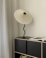 Load image into Gallery viewer, A modern Seashell Table Lamp with a pleated shade and a curvy black base placed on a black and wood cabinet near white curtains, accompanied by an open book.
