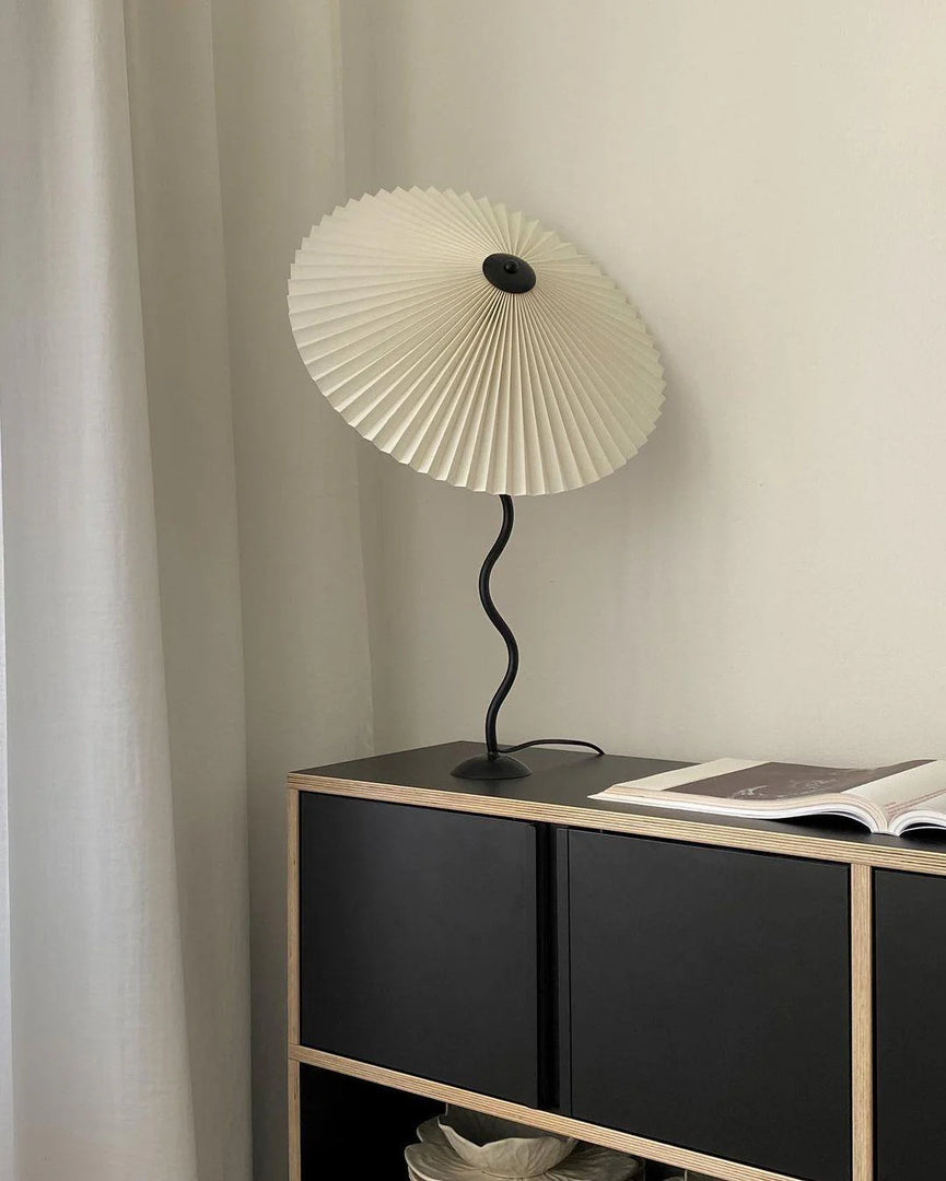 A modern Seashell Table Lamp with a pleated shade and a curvy black base placed on a black and wood cabinet near white curtains, accompanied by an open book.