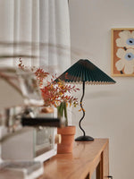 Load image into Gallery viewer, A cozy interior featuring a dark green Seashell Table Lamp with a pleated shade on a wooden table, surrounded by potted plants, autumnal foliage, and a coffee machine, with a floral artwork on the wall.
