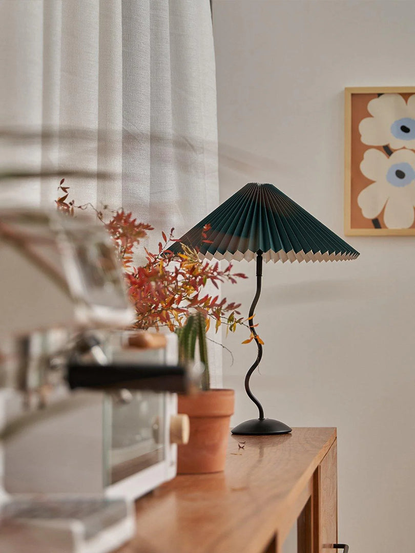 A cozy interior featuring a dark green Seashell Table Lamp with a pleated shade on a wooden table, surrounded by potted plants, autumnal foliage, and a coffee machine, with a floral artwork on the wall.