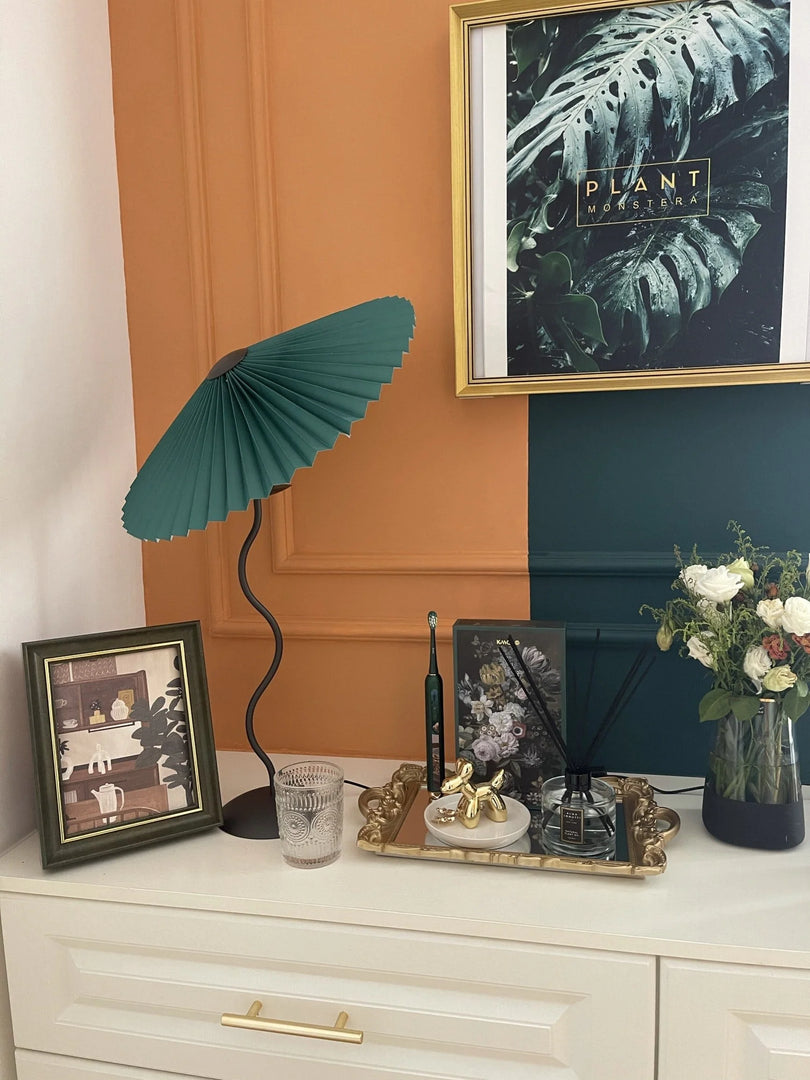 A decorative setup featuring a green Seashell Table Lamp with a pleated shade, a gold tray holding accessories, a vase with fresh flowers, and framed artwork on a white dresser against a dual-tone orange and teal wall.