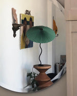 Load image into Gallery viewer, A reflection of a green Seashell Table Lamp with a pleated shade and curvy black base in a circular mirror, placed on a terracotta-style pedestal with artwork and decor visible in the background.
