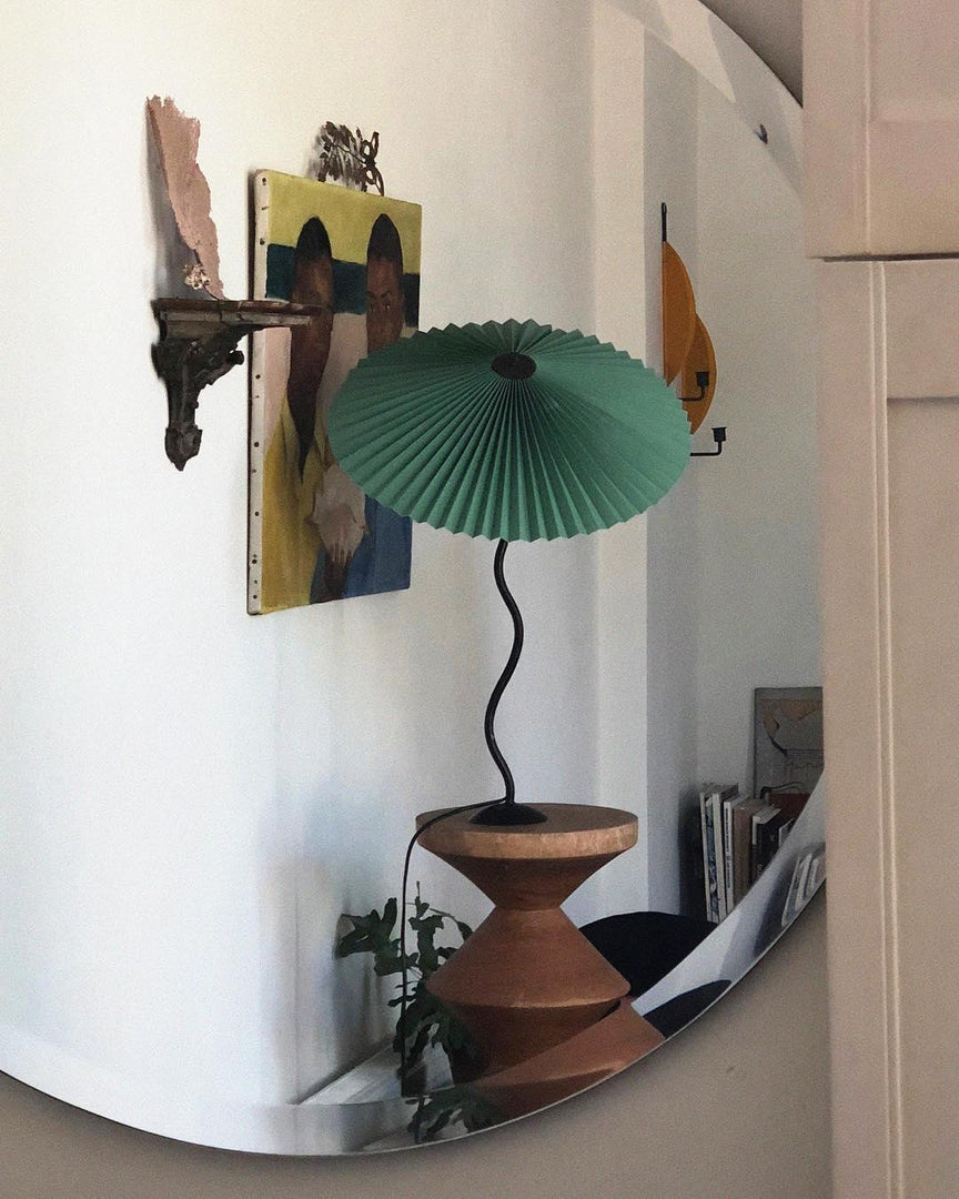 A reflection of a green Seashell Table Lamp with a pleated shade and curvy black base in a circular mirror, placed on a terracotta-style pedestal with artwork and decor visible in the background.