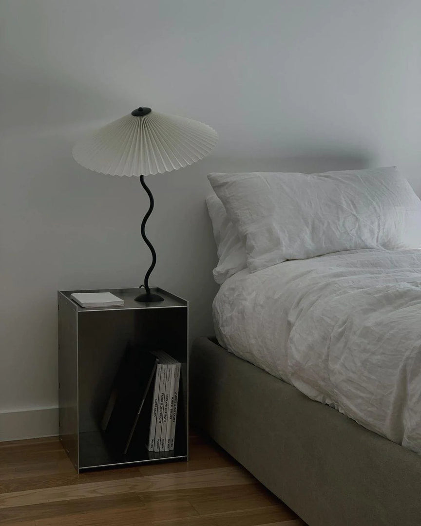 The Seashell table lamp with a pleated white shade and wavy black stand, placed on a sleek metallic side table next to a minimalist bed with crisp white linens, creating a modern and serene bedroom setting.