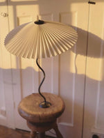 Load image into Gallery viewer, The Seashell table lamp with a pleated white shade and wavy black stand, placed on a rustic wooden stool, casting warm light and shadows against a classic paneled door for a cozy and inviting setting.
