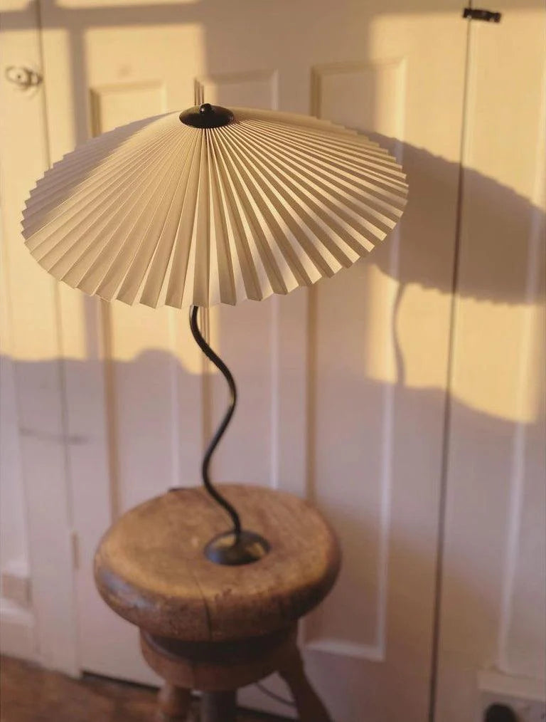 The Seashell table lamp with a pleated white shade and wavy black stand, placed on a rustic wooden stool, casting warm light and shadows against a classic paneled door for a cozy and inviting setting.