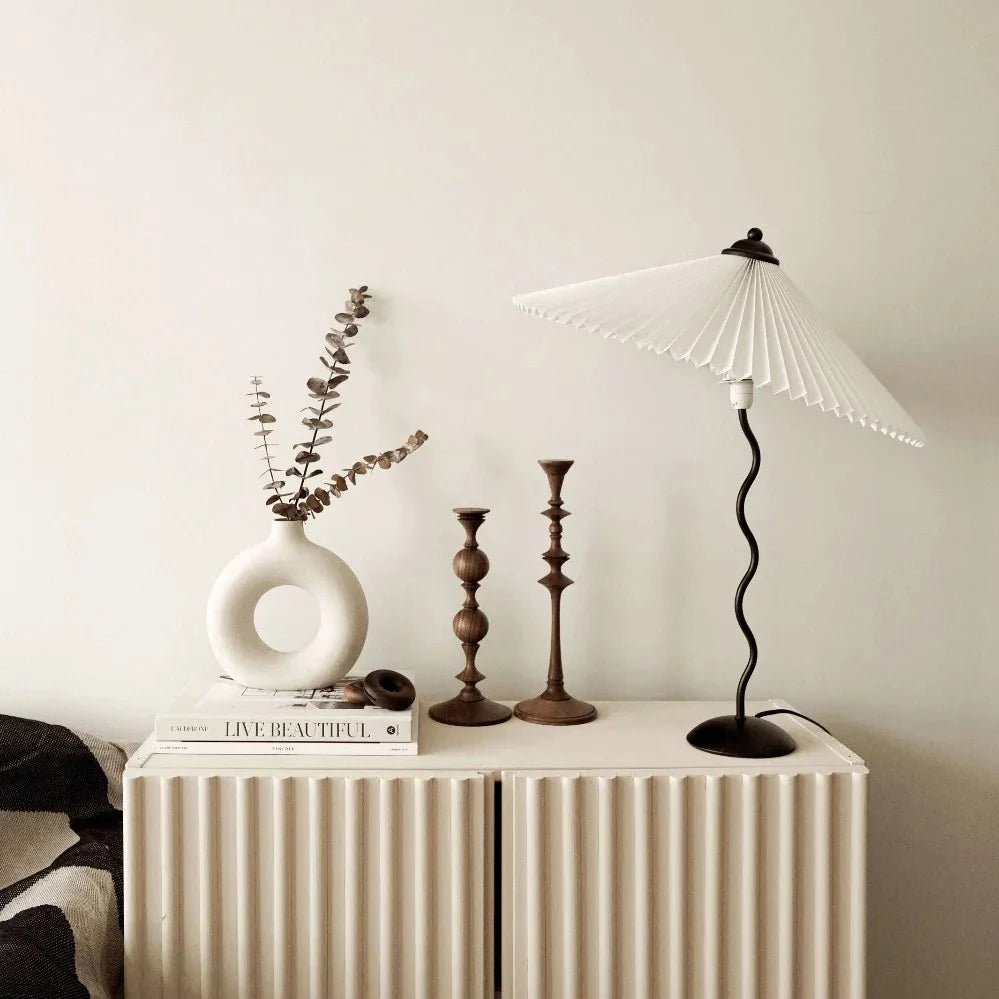 A minimalist tabletop decor setup featuring the Seashell Table Lamp with a pleated shade, a sculptural vase with dried branches, and vintage brass candlesticks on a textured cabinet.