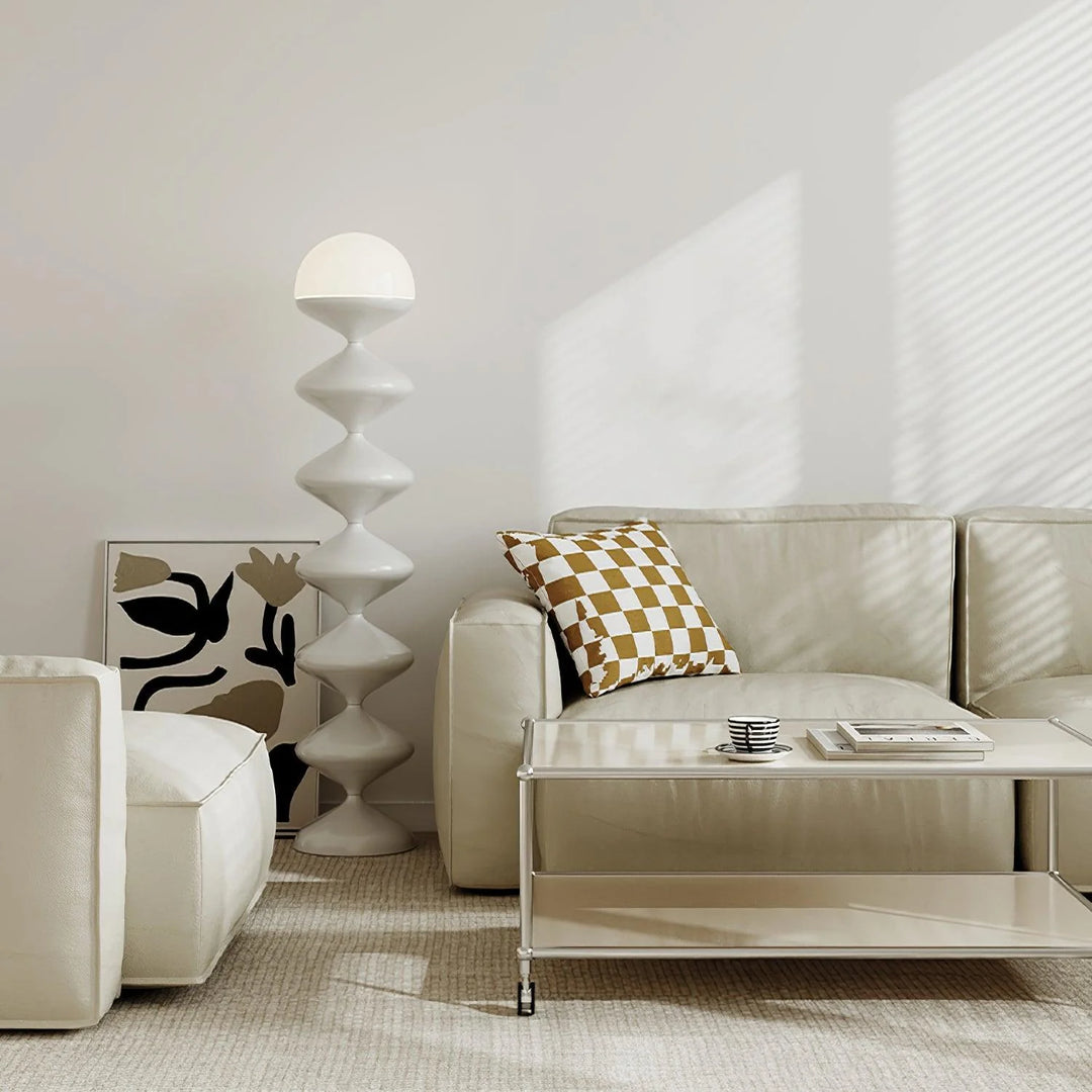 A serene living space highlighted by a Serene White Spiral Lamp with a unique twisting base and spherical top, complementing the off-white couch and matching poufs. The decor is accented by a geometric cushion and a minimalist side table, all bathed in soft, natural light filtering through sheer curtains.