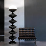 Load image into Gallery viewer, Elegant interior featuring a Sleek Black Gourd Lamp with a vertical stack of glossy black orbs and a contrasting white top, next to a classic brown leather chair against a dark gray wall, showcasing a modern and luxurious design aesthetic.
