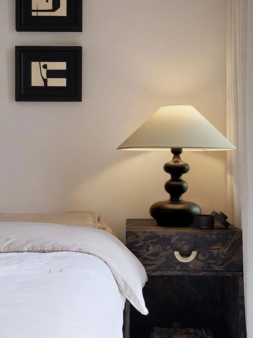 A sophisticated bedroom scene featuring a Gourd Lamp with a dark, elegantly shaped wooden base and a bright white lampshade, placed on a uniquely patterned bedside table. The warm light casts a gentle glow against a neutral wall, complemented by modern black-framed artworks, enhancing the refined and tranquil ambiance of the bedroom.