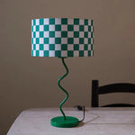 Load image into Gallery viewer, Showcasing the Spiraled Green Checker Lamp, this image presents a captivating green and white checkered lampshade mounted on an elegantly twisted green base. The lamp’s fresh, modern design and vibrant colors make it a distinctive accent piece, positioned on a plain table against a subtle backdrop, emphasizing its quirky and stylish character.
