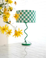 Load image into Gallery viewer, Capturing the essence of spring, this image features the Spring-Inspired Checker Lamp with a vibrant green and white checkered shade on a whimsically spiraled base. The fresh, bright setting is enhanced by the presence of wild yellow flowers in the foreground, placed on a pristine white marble surface, emphasizing a cheerful and lively atmosphere perfect for a modern, nature-inspired decor.
