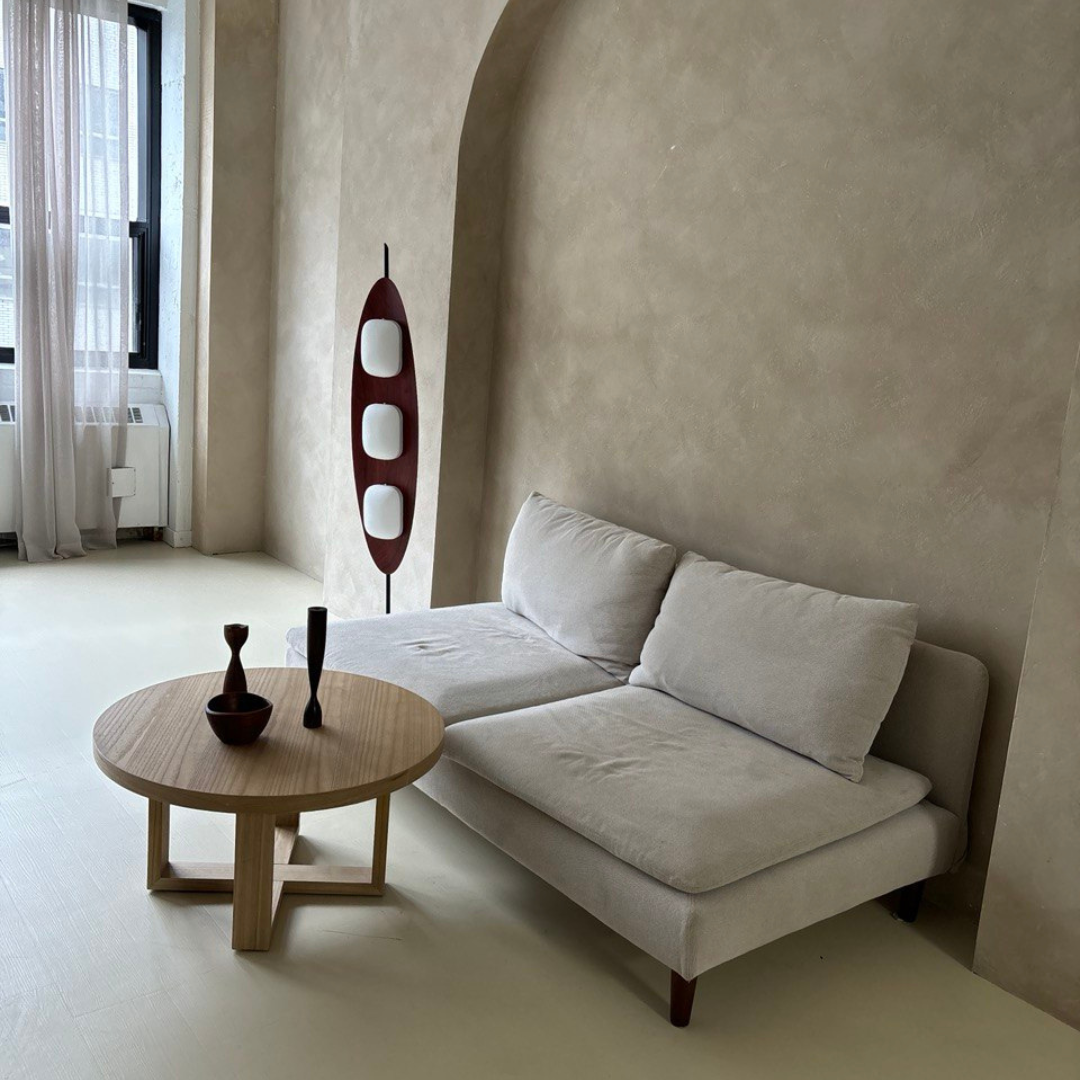 A Surfboard Floor Lamp with a wooden frame and glowing panels positioned next to a neutral-toned sofa and a minimalist round wooden coffee table, set in a bright room with an arched wall and large windows with sheer curtains.