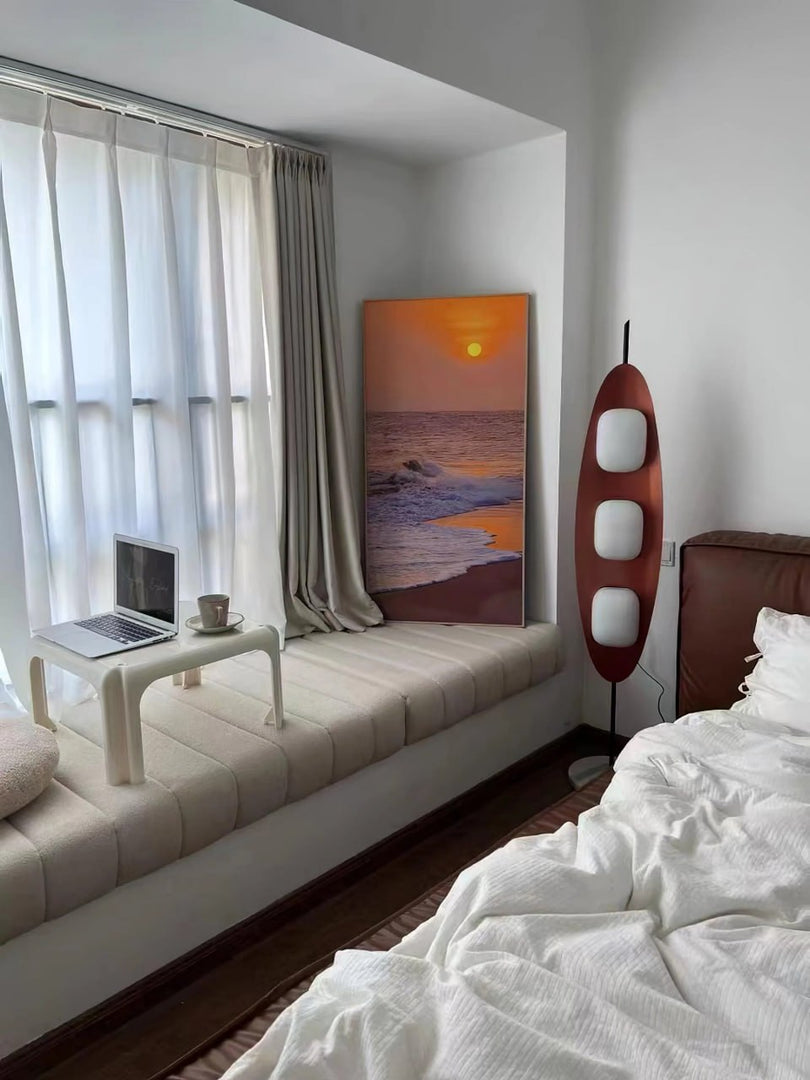 A Surfboard Floor Lamp with a sleek wooden design and glowing panels placed beside a cozy window seat with a laptop and coffee cup, a sunset painting leaning against the wall, and a neatly styled bed in a serene bedroom setting.