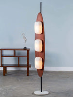 Load image into Gallery viewer, A stylish surfboard-shaped floor lamp with three frosted glass bulbs on a wood frame and a round marble base, placed next to a mid-century modern console table with a decorative bird sculpture, against a light blue wall.
