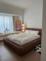 Load image into Gallery viewer, A Surfboard Floor Lamp with a wooden frame and glowing panels positioned beside a luxurious brown leather bed with white bedding, a window seat with a laptop and coffee cup, and a sunset painting in a bright, modern bedroom.
