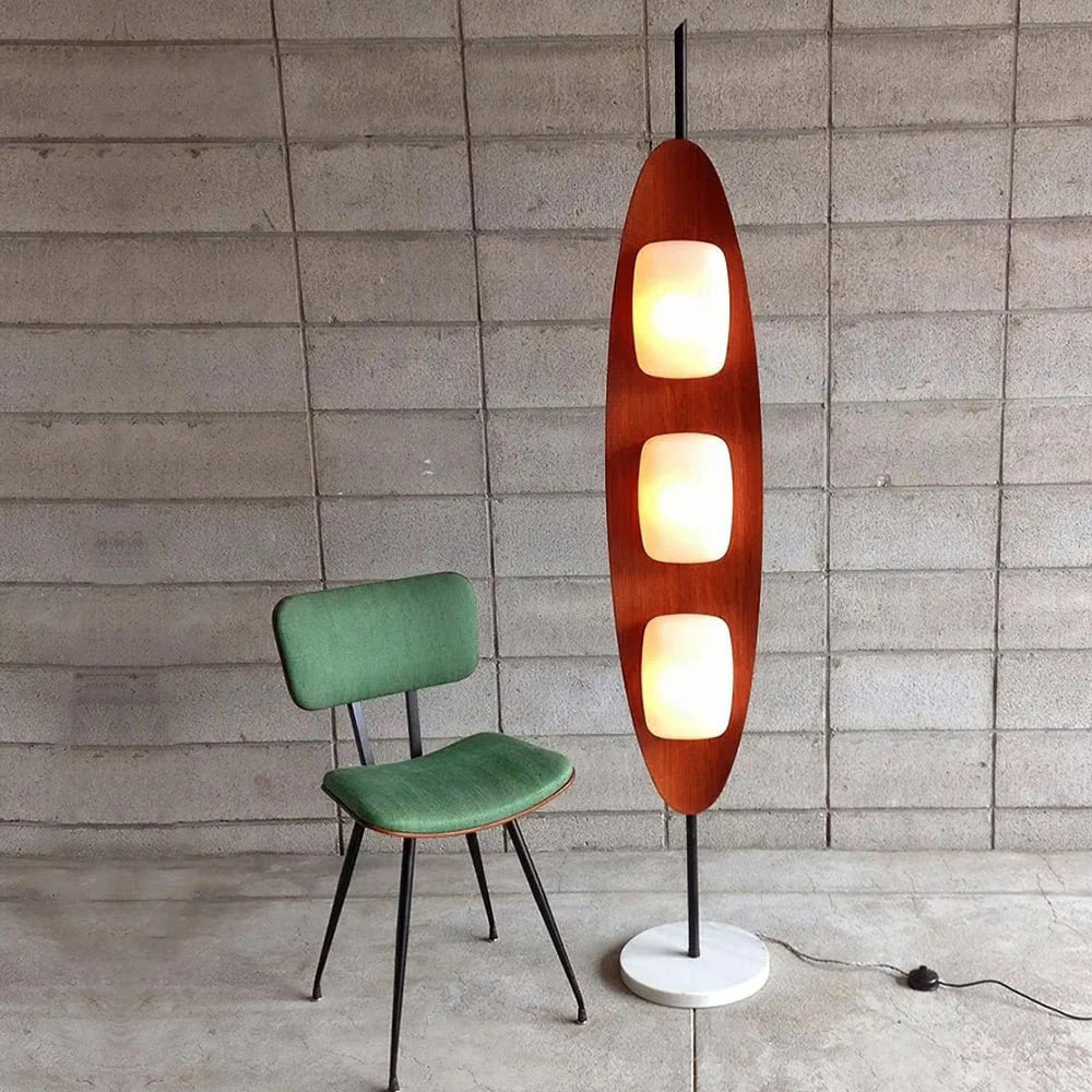 A Surfboard Floor Lamp with a mid-century design featuring a wooden frame, glowing panels, and a marble base, styled beside a retro green upholstered chair against a concrete block wall in a vintage-inspired setting.