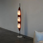 Load image into Gallery viewer, A tall, modern surfboard-shaped floor lamp with three illuminated frosted glass bulbs, resting on a sleek black metal stand and a round white base, placed against a minimalist wall.
