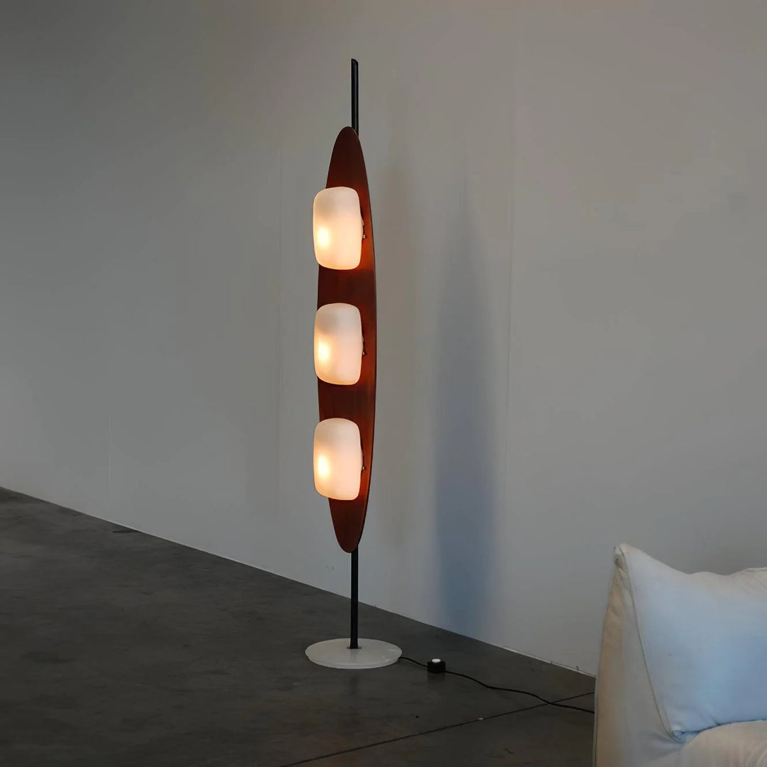 A tall, modern surfboard-shaped floor lamp with three illuminated frosted glass bulbs, resting on a sleek black metal stand and a round white base, placed against a minimalist wall.