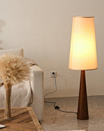 Load image into Gallery viewer, Tall wooden floor lamp with tapered base and cream fabric shade creating warm ambient lighting in a cozy corner with textured wall, beige sofa and decorative pampas grass arrangement
