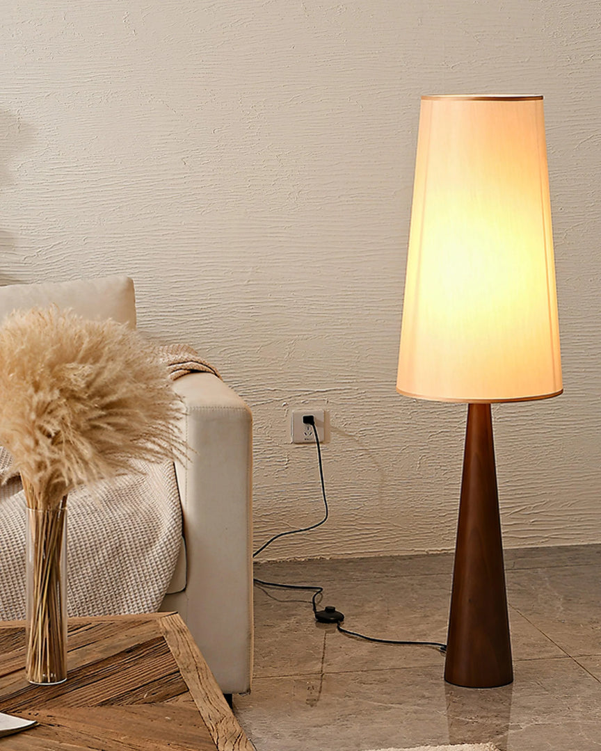 Tall wooden floor lamp with tapered base and cream fabric shade creating warm ambient lighting in a cozy corner with textured wall, beige sofa and decorative pampas grass arrangement