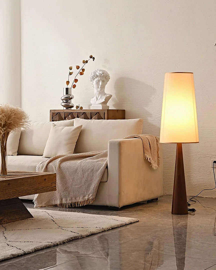 Stylish tapered floor lamp with wooden base and soft glowing shade showcased in neutral living room with cream sofa, geometric rug, and decorative accents including classical bust and dried botanicals