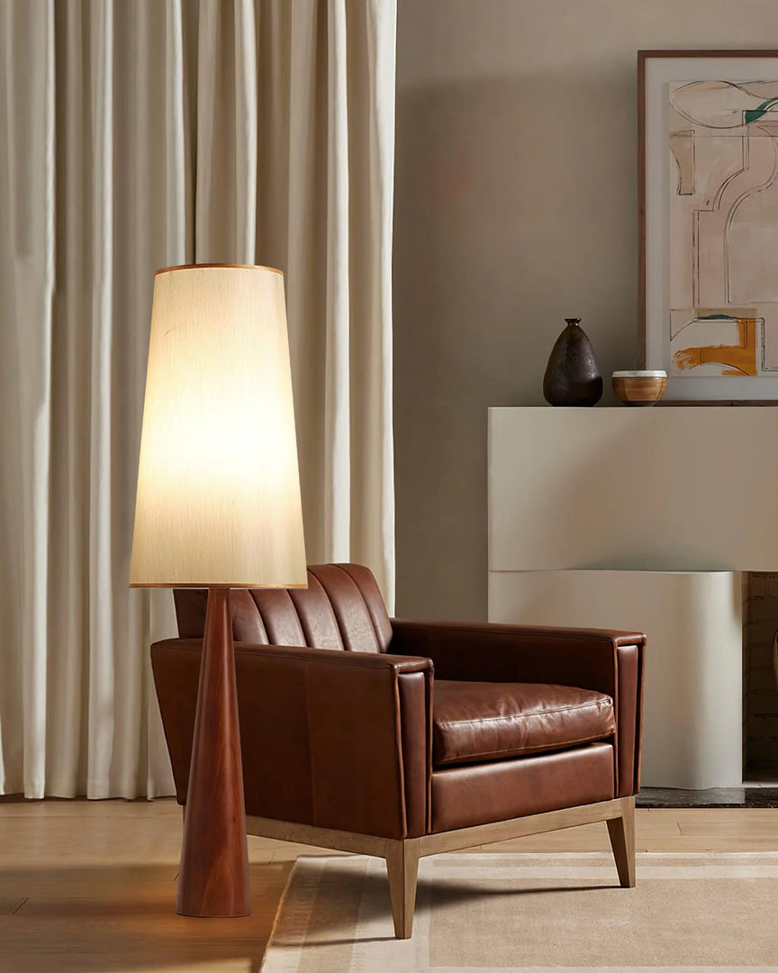 Contemporary floor lamp with conical wooden base and warm white fabric shade paired with brown leather armchair in minimal living room setting featuring modern decor and neutral curtains