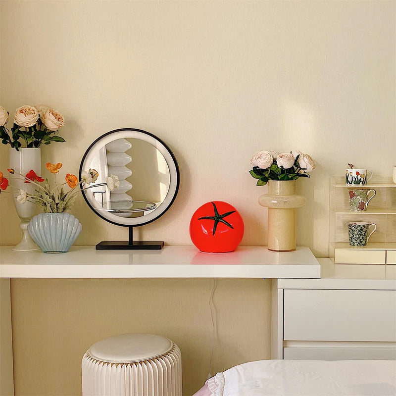 A tomato-shaped table lamp with a glossy red finish and green stem design, placed on a white vanity table surrounded by decorative items, including vases with flowers, a circular mirror, and stacked teacups.