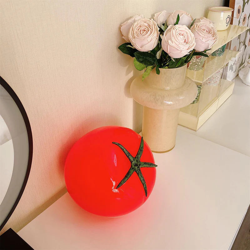 A tomato-shaped table lamp with a glossy red finish and green stem design, placed on a white surface next to a beige vase holding pink roses.