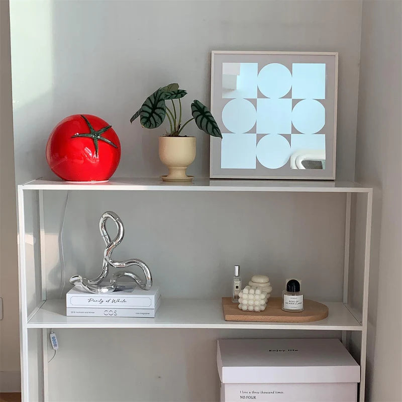 A tomato-shaped table lamp with a glossy red finish and green stem design, placed on a white shelf alongside decorative items, including a potted plant, abstract artwork, and modern decor pieces.