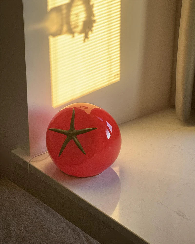 A tomato-shaped table lamp with a glossy red finish and a green stem design, placed on a windowsill with soft sunlight streaming through blinds.