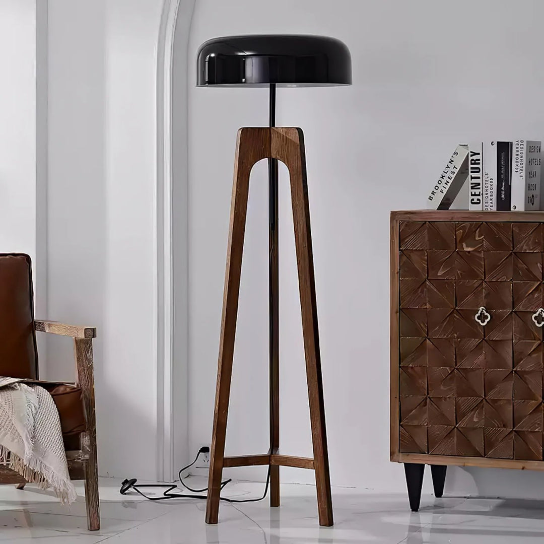 A modern floor lamp with a wooden tripod base and black dome-shaped shade, placed in a bright room next to a vintage wooden cabinet and a rustic armchair draped with a blanket.