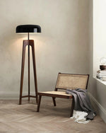 Load image into Gallery viewer, A modern floor lamp with a wooden tripod base and black dome-shaped shade, placed in a cozy corner with a woven chair, soft blanket, and a stack of books on a windowsill.
