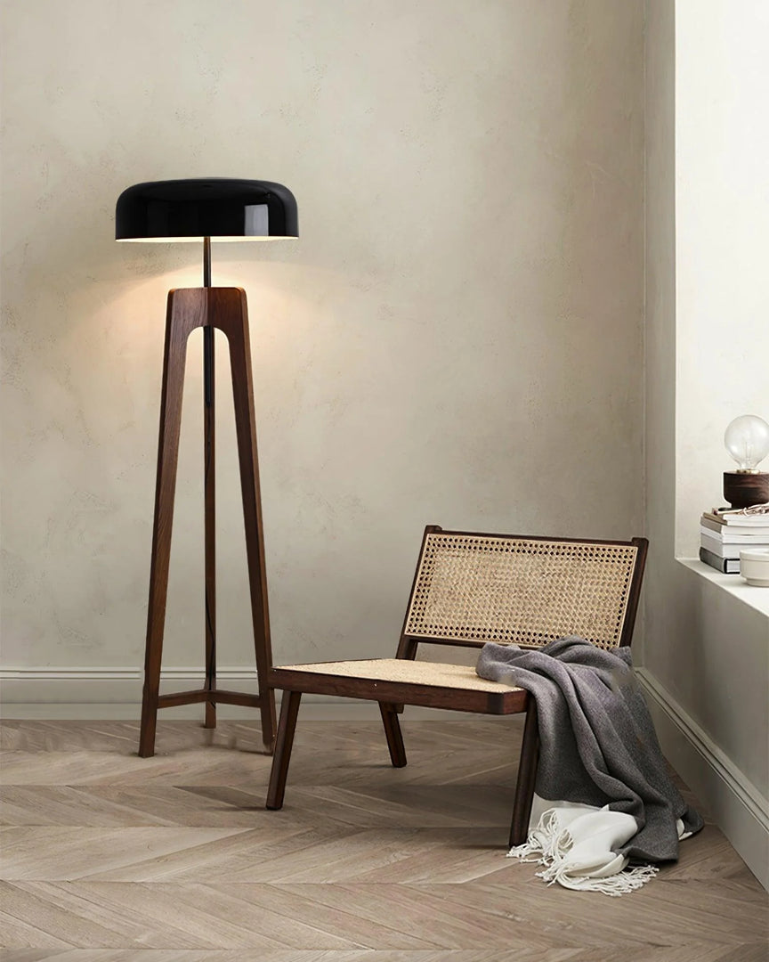 A modern floor lamp with a wooden tripod base and black dome-shaped shade, placed in a cozy corner with a woven chair, soft blanket, and a stack of books on a windowsill.