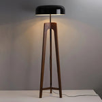 Load image into Gallery viewer, A sleek floor lamp with a wooden tripod base and a black dome-shaped shade, emitting warm light against a neutral gray background, highlighting its modern and minimalist design.
