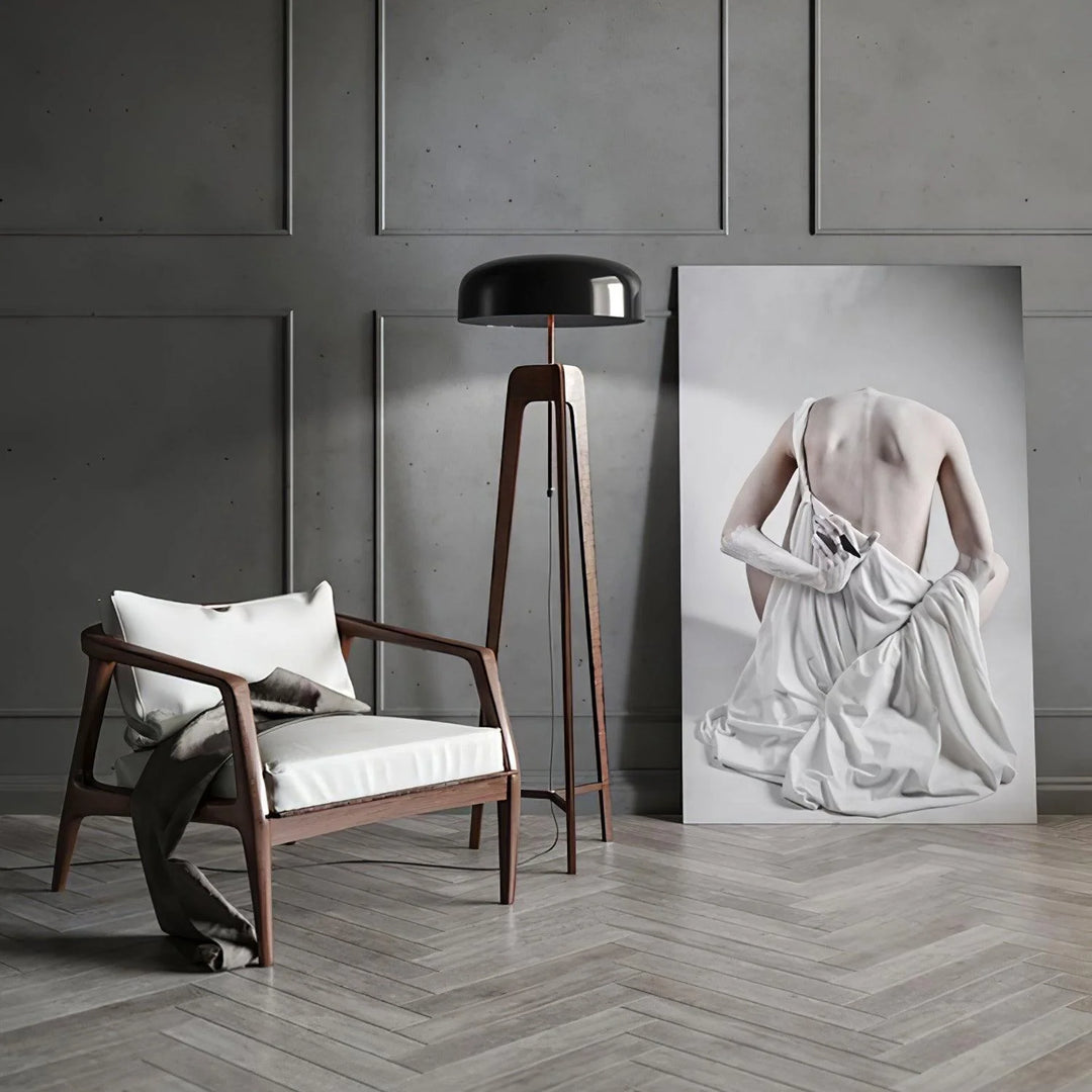 A stylish floor lamp with a wooden tripod base and black dome-shaped shade, set in a modern interior with a wooden armchair, neutral cushions, and an artistic portrait leaning against a gray paneled wall.