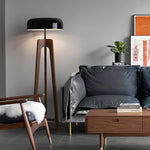 Load image into Gallery viewer, A modern floor lamp with a wooden tripod base and black dome-shaped shade, accenting a stylish living room with a leather sofa, wooden armchair, coffee table, and contemporary decor.
