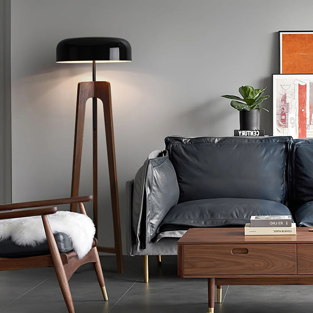 A modern floor lamp with a wooden tripod base and black dome-shaped shade, accenting a stylish living room with a leather sofa, wooden armchair, coffee table, and contemporary decor.