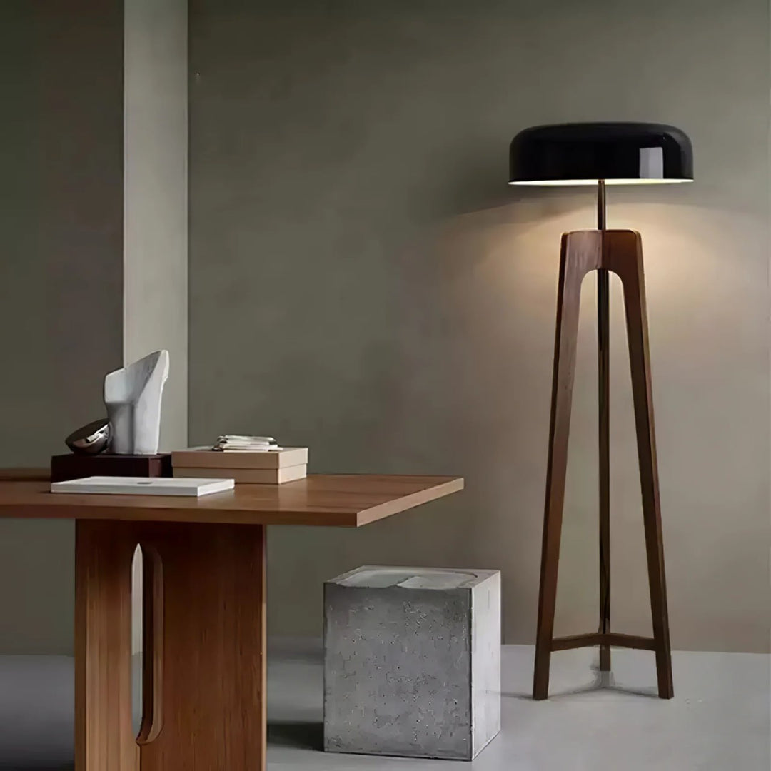 A contemporary floor lamp with a wooden tripod base and a black dome-shaped shade, illuminating a minimalist room featuring a wooden table, a concrete stool, and sculptural decor.