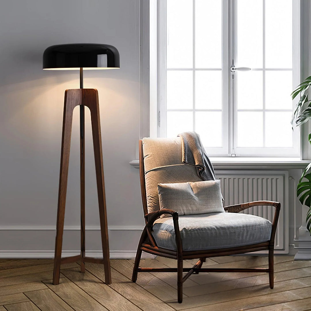 A modern floor lamp with a wooden tripod base and black dome-shaped shade, lighting up a cozy reading nook featuring a cushioned armchair and a large window in a bright, minimalist interior.