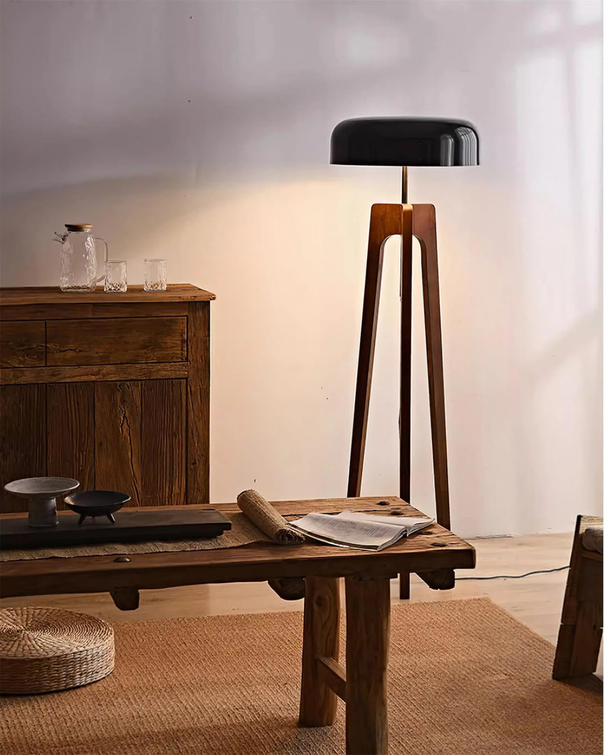 A contemporary floor lamp with a wooden tripod base and black dome-shaped shade, illuminating a rustic interior with wooden furniture, a woven rug, and minimal decor.