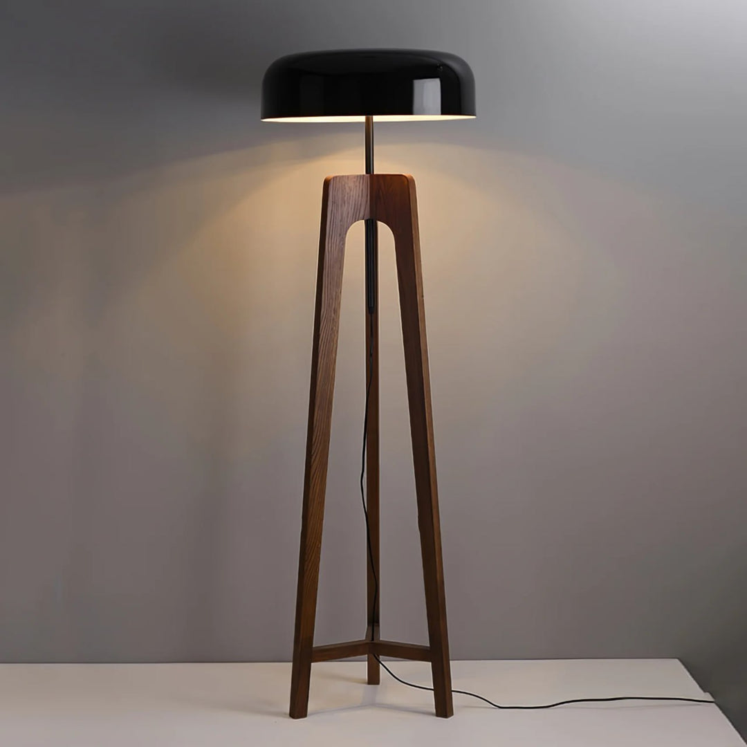 A modern floor lamp with a wooden tripod base and a black dome-shaped shade, emitting warm light against a neutral gray background, showcasing its clean and minimalist design.