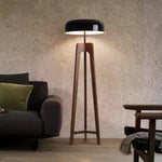 Load image into Gallery viewer, A modern floor lamp with a wooden tripod base and a black dome-shaped shade, emitting warm light in a cozy living room setting with a sofa and armchair nearby.
