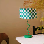 Load image into Gallery viewer, This image features the Twisted Checkerboard Design Lamp, which stands out with its vibrant green and white checkered lampshade and uniquely spiraled base. The lamp is elegantly placed on a white table, providing a pop of color and whimsical style against a neutral backdrop, complemented by soft, natural elements like the delicate, dried flowers in the background. This setup exemplifies a playful yet stylish modern home decor.
