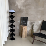 Load image into Gallery viewer, Modern interior showcasing a Unique Gourd Floor Lamp with black stacked gourd base and a white top, positioned next to a wooden stump table and black chair, against a textured grey wall in a minimalist setting.
