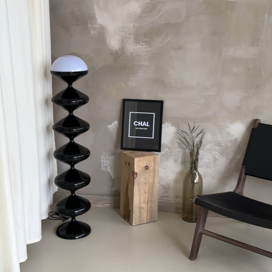 Modern interior showcasing a Unique Gourd Floor Lamp with black stacked gourd base and a white top, positioned next to a wooden stump table and black chair, against a textured grey wall in a minimalist setting.