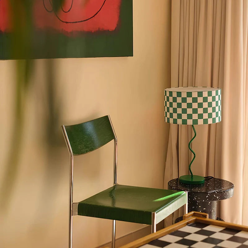 A vibrant setting accentuated by the Chessboard Lamp with its green and white checkered shade and whimsically twisted green base, perfectly placed in a creative corner. The lamp is accompanied by a green chair and a speckled side table, under a bold abstract painting, illustrating a lively and artistic interior scene. The coordinated color scheme and playful design elements create a dynamic and visually appealing space.